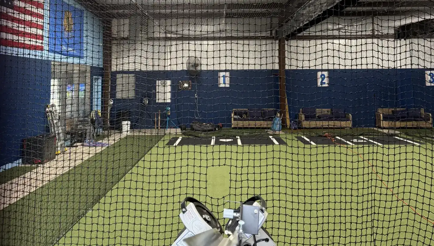 View through the batting cage net past a pitching machine to numbered hitting lanes at Home Plate Sports